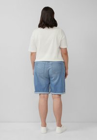 Person, der einer hellgrauen Wand gegenübersteht, trägt ein weißes T-Shirt, hochgekrempelte hellblaue Jeansshorts und weiße Sneakers.