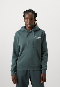 Žalias gobtuvė su raišteliu, kengūros kišenė ir baltos spalvos logotipas. Pagaminta iš švelnios medžiagos, laisvo kirpimo su numegztais rankogaliais.