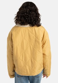 Giacca gialla trapuntata con un morbido colletto in shearling beige. Presenta una forma oversize e una texture liscia. Vista posteriore, orlo curvato.