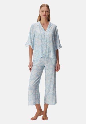 Femme debout pieds nus portant un ensemble pyjama ample bleu clair à motif cachemire, avec un haut à manches courtes boutonné et un pantalon large et court.