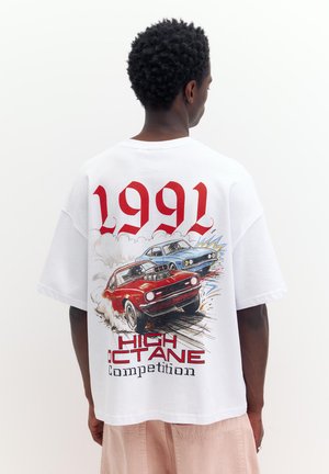 Férfi fehér, túlméretezett pólót visel, amelyen piros és kék izomautók versenyeznek és a „1991 High Octane Competition” felirat szerepel a háton.