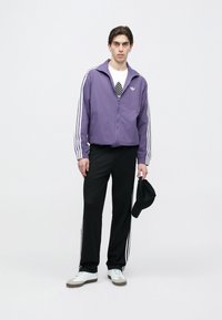 Lillaka tõmblukuga jakk valgete triipudega, mustad dressipüksid ja valged tossud. Riietusele lisandub must nokamüts käes.