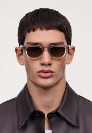 Des lunettes de soleil avec une monture grise translucide et des verres foncés, présentant un design rectangulaire. Portées par une personne en veste en cuir noir.