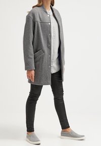 Cappotto grigio in misto lana con chiusura a bottoni, tasche laterali, colletto a coste e vestibilità rilassata, indossato sopra una camicia chiara e pantaloni neri.