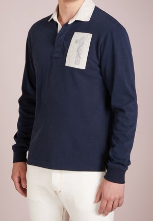 Granatowa koszulka polo z długim rękawem i białym kołnierzykiem, z naszywką na piersi z numerem "7" w kolorze szarym. Materiał bawełniany.