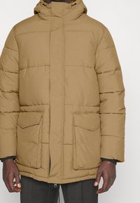 Tan pufferjakke med høj krave, to frontlommer og quiltet design. Fremstillet af letvægts, vandafvisende materiale.