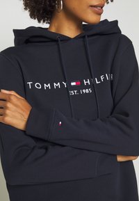 Femme portant un hoodie marine avec le texte et le logo "Tommy Hilfiger Est. 1985", les bras croisés, montrant une partie de son visage et ses cheveux bouclés.