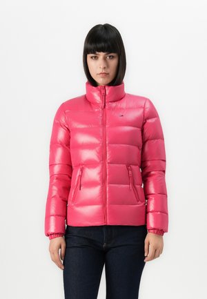 Vrouw in een felroze gewatteerde pufferjack met ritszakken, staat tegen een effen witte achtergrond.