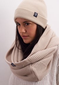 Beige knit beanie and matching infinity scarf, featuring ribbed and textured patterns, with a small logo tag on each item.