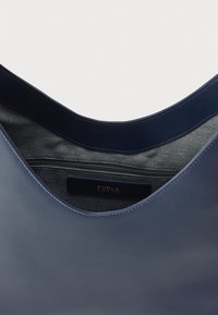 Modrá kožená kabelka s hladkou textúrou, zaobleným otvorom, sivou vnútornou podšívkou a zlatým vyrazeným štítkom značky vo vnútri.