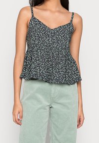 Černý peplum top s bílým a zeleným květinovým vzorem, výstřihem do V a tenkými ramínky, v kombinaci se světle zelenými manšestrovými kalhotami.
