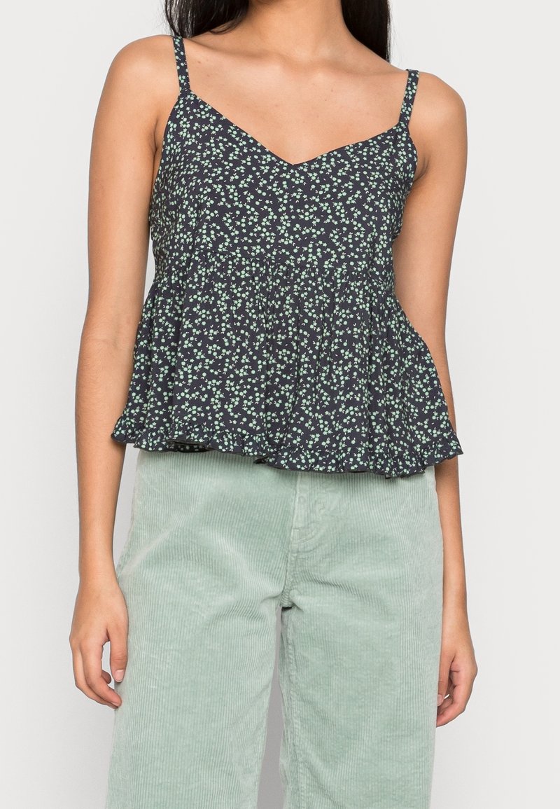 Černý peplum top s bílým a zeleným květinovým vzorem, výstřihem do V a tenkými ramínky, v kombinaci se světle zelenými manšestrovými kalhotami.