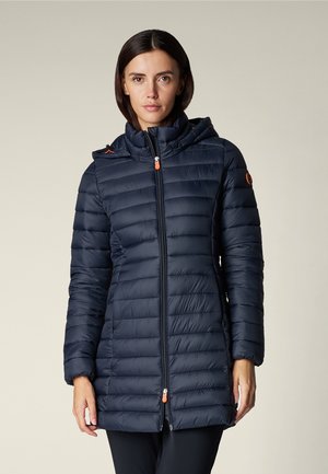 Donna che indossa una giacca imbottita trapuntata lunga blu navy con cappuccio e cerniera frontale, in piedi contro uno sfondo semplice.