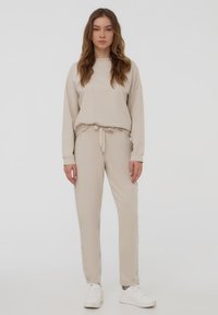 Felpa beige e pantaloni jogger coordinati realizzati in tessuto morbido. La felpa ha un collo rotondo e spalle scese; i pantaloni jogger presentano una vita con cordoncino.