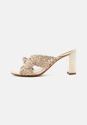 Mule beige con tacco alto, cinturini larghi intrecciati e tacco spesso, design con punta aperta su sfondo bianco neutro.