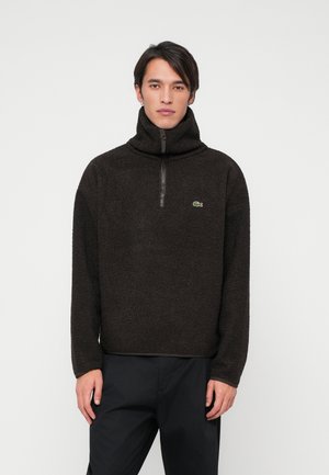 Fleece-Pullover in Dunkelbraun, mit hohem Kragen und halbem Reißverschluss. Verfügt über eine strukturierte Oberfläche und ein kleines grünes Logo auf der Brust.