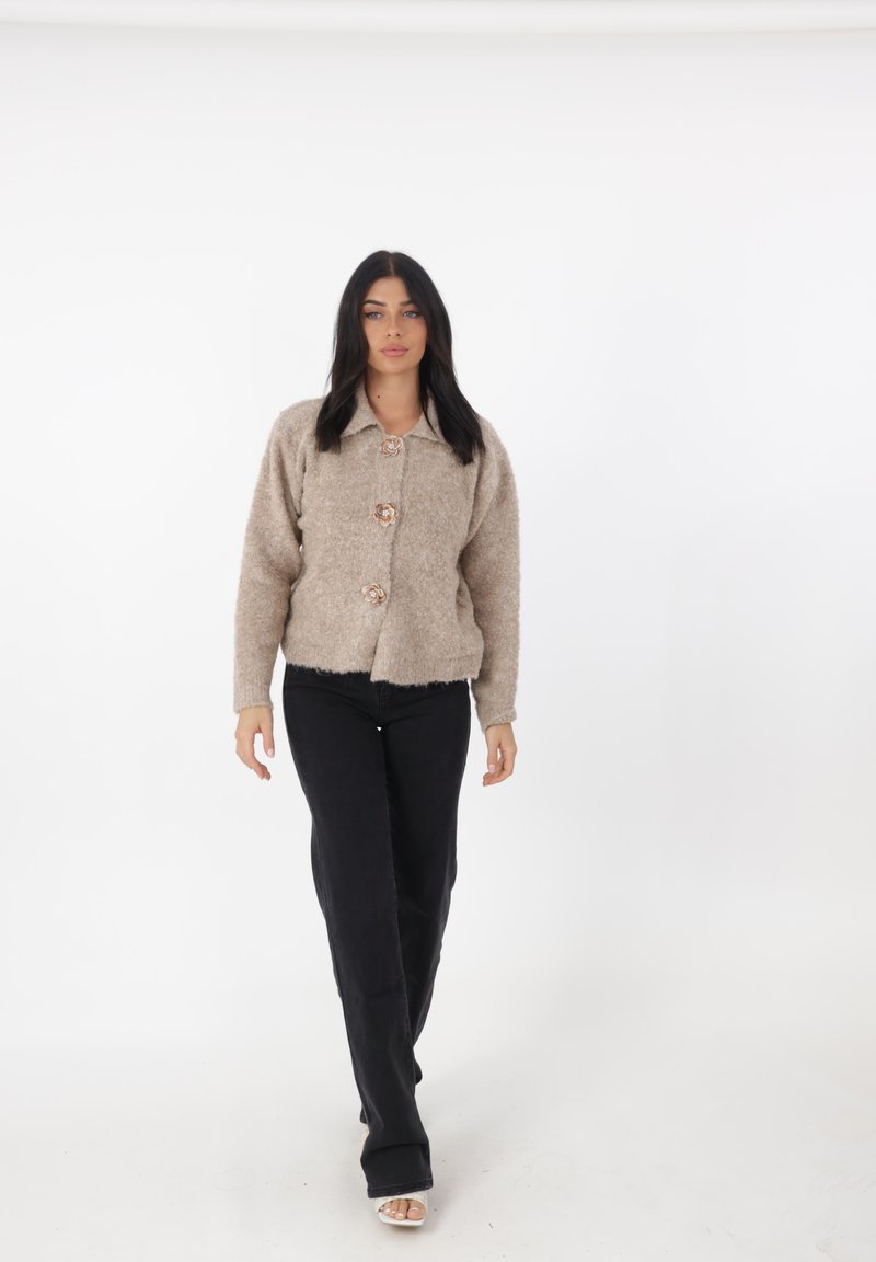 Maglione beige peloso con grandi bottoni, design con colletto e maniche lunghe. Pantaloni neri a gamba larga con taglio dritto. Sfondo neutro.
