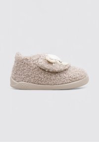 Zapato slip-on de color beige, difuso, con parte superior de textil suave y una solapa decorativa redonda con un diseño de animal sencillo. Suela de goma color crema.
