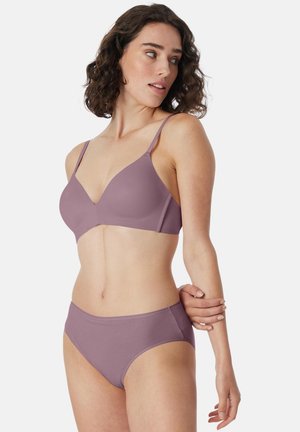 Woman with curly dark hair wearing matching mauve bra and underwear, looking to her left, with arm bent behind her back.