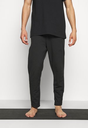 Homme debout pieds nus sur un tapis noir, portant un pantalon décontracté noir et une chemise noire, sur un fond blanc uni.