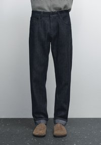 Mörk denimjeans med avslappnad passform, har knäppning med knapp, femfickorsdesign och uppvikta benslut, i kombination med bruna skor.