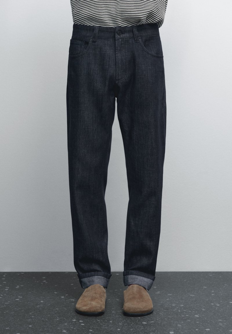 Mörk denimjeans med avslappnad passform, har knäppning med knapp, femfickorsdesign och uppvikta benslut, i kombination med bruna skor.