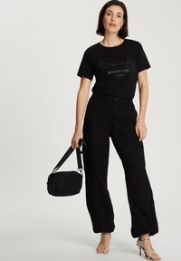 Czarny t-shirt z napisem "Tworzenie Wspomnień Każdego Weekendu", w połączeniu z wysokimi czarnymi szerokimi spodniami, srebrnymi sandałami na paski i małą czarną torbą.