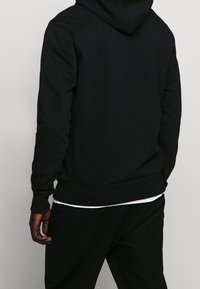 Černá mikina s kapucí s žebrovanými manžetami a lemem, vyrobená z měkké látky; nošena přes bílé tričko, s jednoduchým designem a volným střihem.