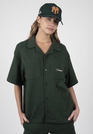 Mujer con camisa de manga corta abotonada de color verde oscuro y gorra a juego con logo naranja de NY, de pie con las manos en los bolsillos sobre fondo blanco.