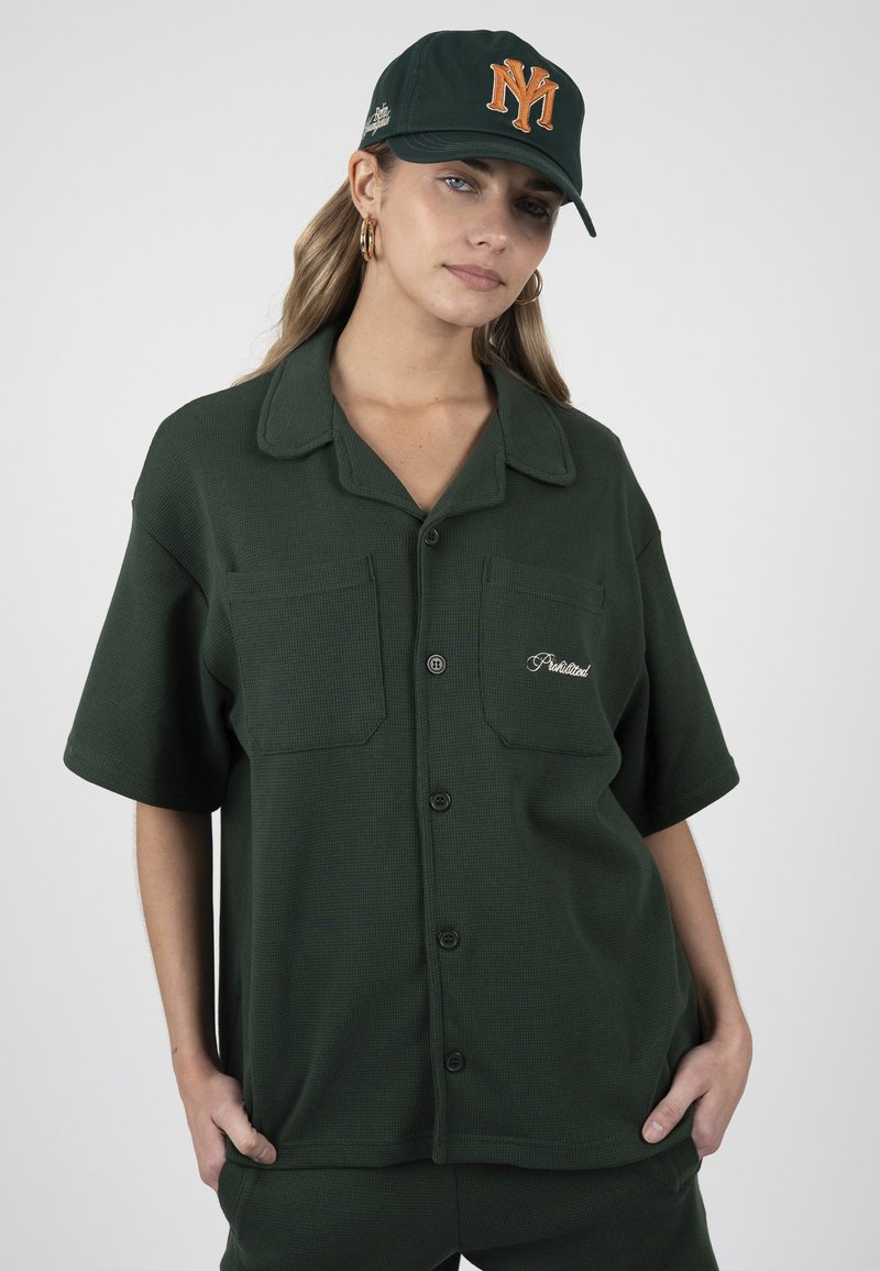 Femme portant une chemise boutonnée à manches courtes vert foncé et une casquette assortie avec un logo NY orange, debout les mains dans les poches sur fond blanc.