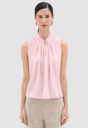 Donna che indossa una blusa senza maniche di colore rosa pallido con pieghe e una spilla di perle al collo, abbinata a pantaloni beige con trama.