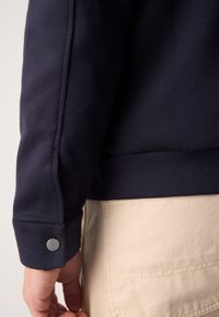 Gros plan sur la main d'une personne près du poignet d'une manche bleu marine avec un bouton argenté, à côté d'une poche de pantalon beige.