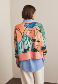 Kleurrijke oversized trui met een bloemmotief in blauw- en groentinten op een perzikkleurige achtergrond. Roze geribde zoom en gelaagd met een lichtblauw overhemd.