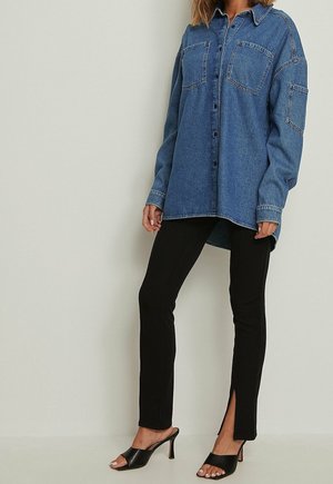 Kvinde iført en blå denim oversize skjorte, sorte tætsiddende bukser med slidser foran, og sorte åbne hælede mule sko, der står op ad en almindelig væg.