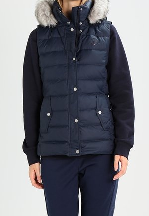 Frau trägt eine dunkelblaue gesteppte ärmellose Weste mit pelzbesetzter Kapuze und Druckknopftaschen über einem langärmeligen Oberteil und Hose.