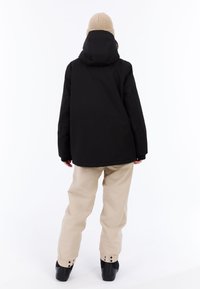 Zwarte geïsoleerde jas met een capuchon, gecombineerd met beige broek en zwarte schoenen. De jas heeft een relaxte pasvorm en elastische manchetten.