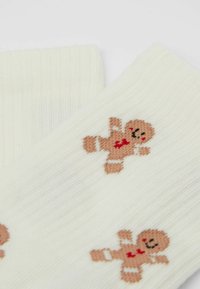 Calzini beige lavorati a maglia con motivi di omini di pan di zenzero in marrone e rosso. Tessuto morbido con polsini a coste, che mettono in risalto i disegni festivi.