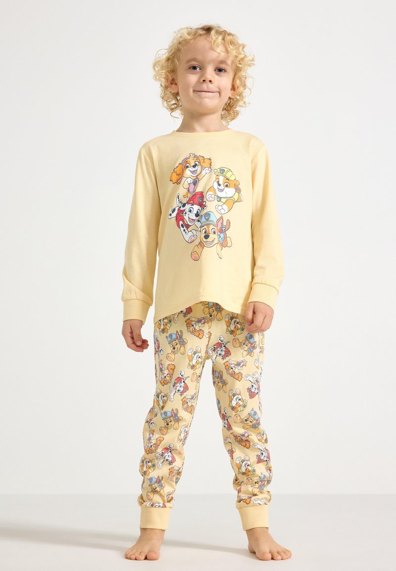 Παιδί με κατσαρά ξανθά μαλλιά που φοράει κίτρινη πυτζάμα με πολύχρωμους κινουμένων σχεδίων χαρακτήρες σκύλων στο πάνω μέρος και στο παντελόνι.