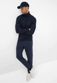 Dunkelblaues Rollkragenoberteil, figurbetontes Design, kombiniert mit dunkelblauen Hosen. Weiße Sneaker mit grauen Akzenten. Model posiert vor einem weißen Hintergrund.