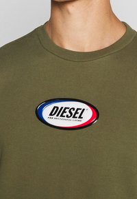 T-shirt verde oliva con un grande patch ovale con la scritta "DIESEL" in nero, contornato di rosso e blu, con il testo che recita "PER UNA VITA DI SUCCESSO."