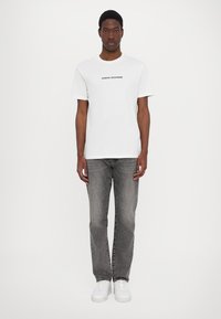 Valge puuvillane t-särk, millel on must "ARMANI EXCHANGE" tekst, koos hallide denim-pükstega ja valgete tossudega. Lihtne disain, mugav istuvus.