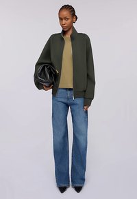 Olivgrön bomberjacka, beige topp, blå jeans med vida ben och svart kuvertväska; modellen står mot en neutral bakgrund och bär spetsiga klackar.