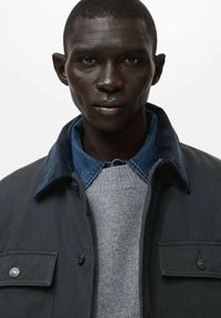 Donkergrijze jas met een corduroy kraag, gelaagd over een lichtgrijze sweater en een blauwe denim shirt. Beschikt over knoopaccenten en een borstzak.