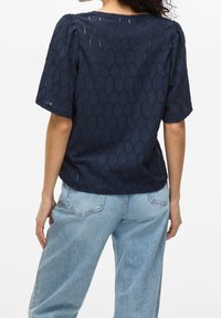 Woman wearing navy blue textured short-sleeve top and light blue high-waisted jeans, shown from the back against a white background.