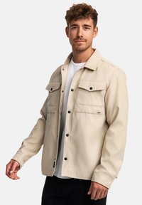 Beige Jacke aus leichtem Stoff mit zwei Brusttaschen, schwarzen Knopfverschlüssen und lässiger Passform, über einem weißen T-Shirt.