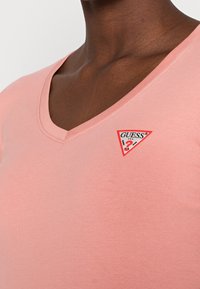 T-shirt rosa a manica corta con scollatura a V, realizzato in un tessuto morbido e con un design aderente. Presenta una toppa con logo a triangolo con dettagli rossi, neri e bianchi.