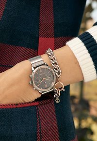 Montre à bracelet en maille argentée avec un cadran sombre et des accents en or rose, accompagnée d'un bracelet en chaîne argentée épaisse avec un charme circulaire.