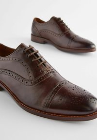 Brown leather dress shoes with perforated detailing, lace-up design, and rounded toe. Textured finish with contrasting stitching and low heel.