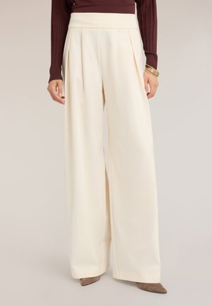 Vrouw draagt crèmekleurige, hoge taille, wijd uitlopende broek met plooien aan de voorkant, gecombineerd met een bruine gebreide top en bruine puntige schoenen.