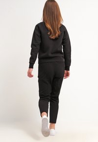 Sort sweatshirt og matchende joggers, ribbede manchetter og talje, glat tekstur, båret med hvide slips-on sko, set fra ryggen.